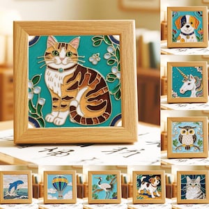 May include: A square wooden frame containing a decorative tile with a colorful stained-glass style illustration of a brown tabby cat sitting against a teal background with white floral accents and green leaves, surrounded by other similar framed tiles.