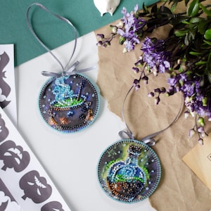 May include: Two round, beaded ornaments with gray ribbons. One features a bubbling cauldron, the other a potion bottle. Both have blue and green accents, with a dark background. The ornaments are likely for Halloween or a similar theme.