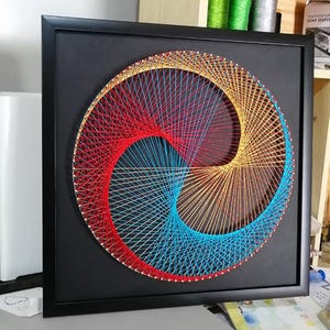 May include: A square black frame containing circular string art on a black background. The design features a swirling pattern created with red, orange, yellow, and blue threads anchored by small silver nails, forming a geometric vortex shape.