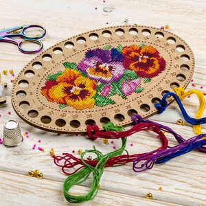 May include: A wooden oval-shaped thread organizer with 32 holes numbered 1 to 32. The organizer has a cross-stitch design of three purple, yellow, and red pansies.  The organizer is surrounded by colorful embroidery floss.
