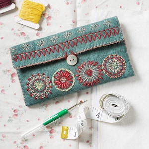 May include: A teal felt needle case with embroidered floral designs in red, pink, and yellow thread. The case has a button closure and is surrounded by a white measuring tape and a needle threader.