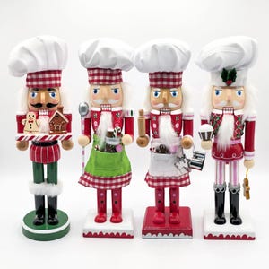 Peut inclure: Quatre figurines de casse-noisettes habillées en chefs, portant chacune une toque blanche avec une bande à carreaux rouges et blancs. Elles présentent divers accessoires de pâtisserie dont un bonhomme en pain d'épices, un rouleau à pâtisserie, un fouet et un cupcake, sur des socles blancs.