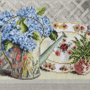 Pu&ograve; includere: Una rappresentazione a punto croce di una natura morta floreale. L'immagine presenta un annaffiatoio rosa pieno di fiori rossi, un annaffiatoio con ortensie blu, un piatto con rose, un piccolo vaso con piante verdi e un secchio verde con fiori viola.