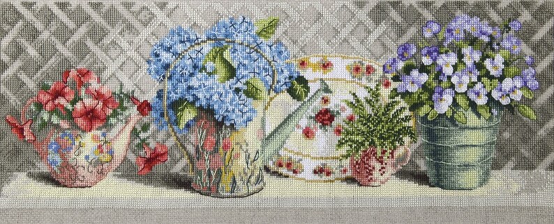 Pu&ograve; includere: Una rappresentazione a punto croce di una natura morta floreale. L'immagine presenta un annaffiatoio rosa pieno di fiori rossi, un annaffiatoio con ortensie blu, un piatto con rose, un piccolo vaso con piante verdi e un secchio verde con fiori viola.