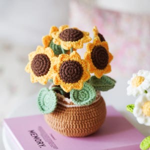 May include: A handcrafted crochet sunflower arrangement in a brown pot. The sunflowers have yellow petals and brown centers, with green leaves. The pot is brown with a white trim. The arrangement is on a pink book.