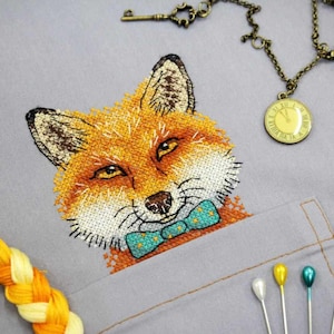 May include: Embroidered fox with a teal bow tie, stitched on a gray fabric background. The fox has orange fur, a white muzzle, and black eyes.