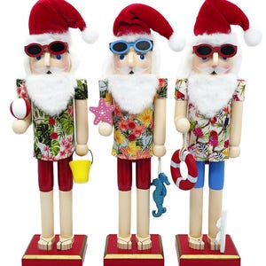 May include: Three nutcracker figures wearing red Santa hats, sunglasses, and tropical print shirts. Each figure has a white beard, red shorts, and sandals. They hold beach accessories including a ball, a yellow bucket, a starfish, a seahorse, and a life ring.