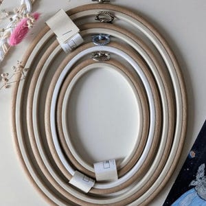 May include: A collection of oval embroidery hoops in varying sizes. The hoops are made of light-colored wood and white plastic, with metal clasps. Some hoops have paper tags attached. The image also includes embroidery floss and a small illustration.