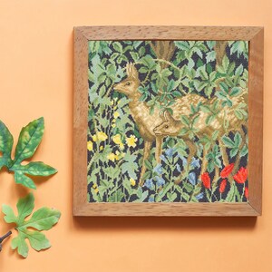 May include: A framed cross-stitch tapestry depicting two deer in a forest setting. The deer are brown and white, and the forest is filled with green leaves and flowers. The tapestry is framed in a natural wood frame.