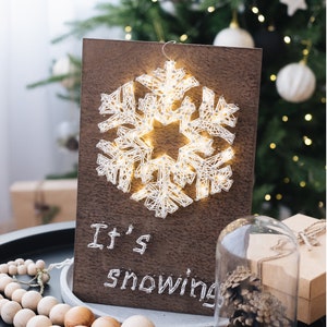 May include: A wooden sign with a white string art snowflake and the text "It's snowing" on a dark brown background. The sign is sitting on a table with a string of wooden beads and a glass cloche with a pine cone inside.
