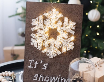 May include: A wooden sign with a white string art snowflake and the text "It's snowing" on a dark brown background. The sign is sitting on a table with a string of wooden beads and a glass cloche with a pine cone inside.