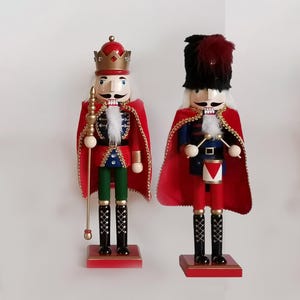 May include: Two decorative wooden nutcrackers. One wears a red and gold crown, red cape, and holds a gold staff. The other wears a black hat with a red plume, a red cape, and holds drumsticks. Both have white hair and mustaches.