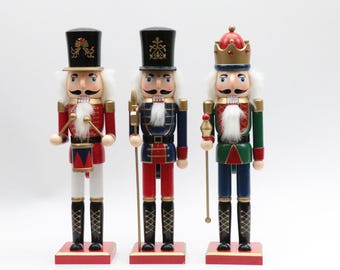 May include: Three wooden nutcracker figurines standing on rectangular bases. The left figure wears a red uniform with a drum, the center figure wears a blue uniform with a spear, and the right figure wears a green uniform with a crown and scepter.