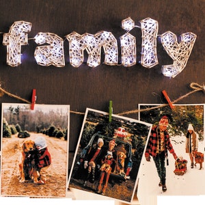 May include: A string art piece spelling "family" with white string wrapped around nails on a brown wooden background. Three photos are hung below with pegs on a twine line. The photos show a family at a Christmas tree farm.