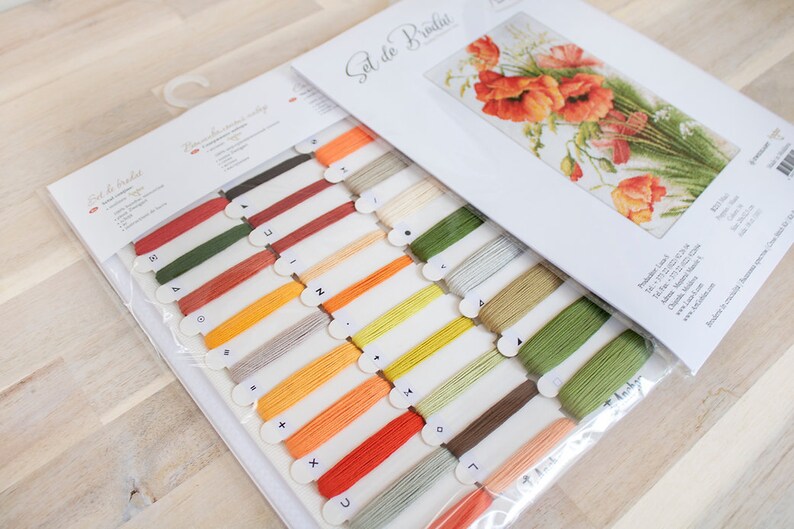 May include: A cross-stitch kit featuring a floral design with red poppies and green foliage. The kit includes a variety of colored embroidery floss and a printed pattern. The packaging displays the text "Set de Broderie".