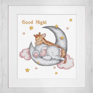 May include: A framed cross-stitch artwork featuring a sleeping elephant and giraffe nestled on a crescent moon. The image includes the words "Good Night" and is surrounded by stars and pink clouds. The artwork is framed in a white frame.