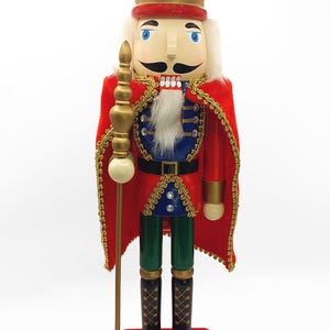 May include: A decorative wooden nutcracker in the form of a king, wearing a red cape with gold trim, a gold crown, and holding a staff. The nutcracker has a white beard, blue eyes, and a black mustache. It stands on a red base.