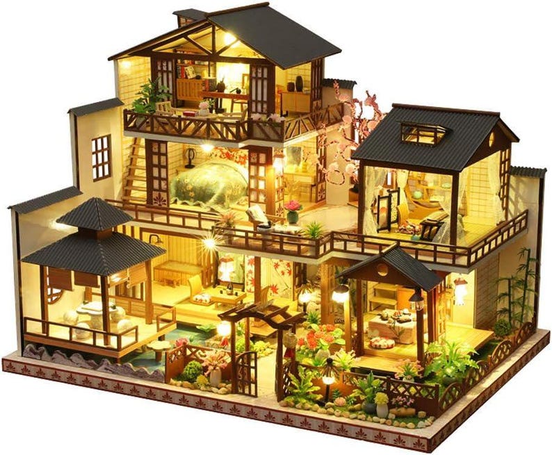 May include: A multi-level dollhouse with a wooden frame and a light brown roof. The dollhouse has a balcony, windows, and a garden with flowers and trees. The dollhouse is lit up with small lights.
