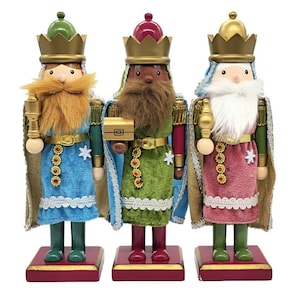May include: Three nutcracker figures standing on square bases, each wearing a gold crown. The left figure wears blue, the centre green, and the right pink. They feature faux fur beards, capes, and carry small gold objects in their hands.