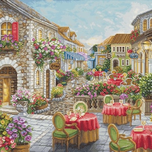 May include: A detailed cross-stitch depicting a European street scene. Stone buildings with red awnings and flower boxes line the cobblestone street. Tables with red tablecloths and green chairs are set for dining. The sky is blue.