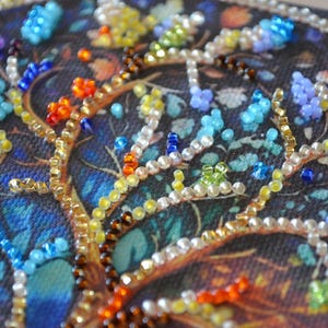 May include: Close-up of a beaded artwork with a tree design. The branches are decorated with gold, brown, and white beads. The leaves and flowers are made of blue, orange, yellow, and green beads, set against a dark blue background.