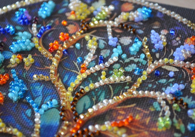 May include: Close-up of a beaded artwork with a tree design. The branches are decorated with gold, brown, and white beads. The leaves and flowers are made of blue, orange, yellow, and green beads, set against a dark blue background.