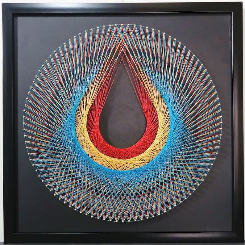 May include: A square black frame containing string art on a black background. The artwork features a circular arrangement of silver nails connected by vibrant red, yellow, and bright blue threads, creating a central teardrop-shaped negative space.