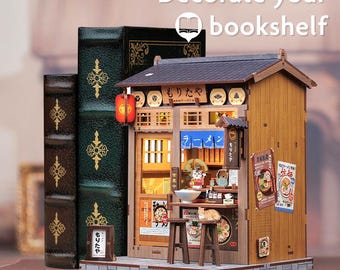 May include: A miniature wooden Japanese ramen shop model with a red lantern, small stools, and posters. The model is placed next to three decorative books. The text "Decorate your bookshelf" is at the top.
