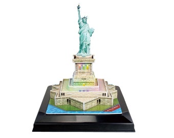 May include: A 3D puzzle model of the Statue of Liberty, featuring a green statue on a stone base with a black display stand. The base has a colorful, rainbow-like design.