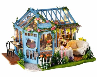 Puede incluir: Una réplica de casa de muñecas en miniatura de una tienda de té azul y blanca con un techo de cristal y un letrero que dice "The Blue Tree English Sweet Shop". La tienda tiene un pequeño patio con una mesa y sillas, y una valla blanca de madera.