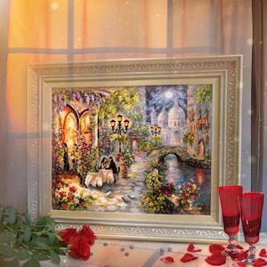 May include: A framed cross-stitch artwork depicting a romantic scene in a European city at night. The artwork features a couple dining at a table outside a building, a bridge over a canal, and a church in the distance. The artwork is framed in a gold-coloured frame.