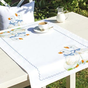 May include: White table runner with a blue embroidered border and a design of three white geese with blue ribbons. The runner is on a white table with a white cup and saucer and a glass pitcher.