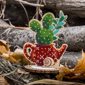 May include: A wooden craft kit featuring a cactus in a teapot with a snail on the side. The cactus is green with white dots and has a small green branch. The teapot is red with white and yellow dots. The snail is brown with a white shell. The kit is on a wooden base.