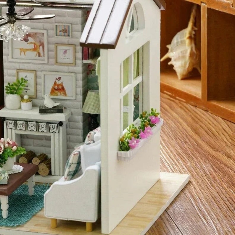 Puede incluir: Un sal&oacute;n de casa de mu&ntilde;ecas en miniatura con un sof&aacute; blanco, una chimenea con una fachada de ladrillo y una ventana con una jardinera. La habitaci&oacute;n est&aacute; decorada con varias im&aacute;genes y una pancarta que dice "Happy".