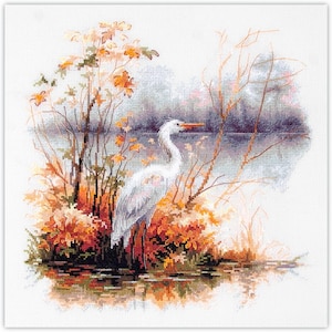 May include: Cross-stitch embroidery of a white heron standing in water, surrounded by autumn foliage. The scene includes trees with orange and yellow leaves, reflecting in the water. The heron has a long neck and orange beak.
