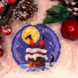 May include: A purple and blue Christmas ornament with a cross-stitch design of Santa Claus climbing down a chimney with a black cat watching from below. The ornament is surrounded by a snowy background.