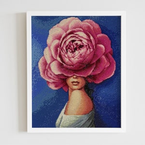 May include: A woman's face is obscured by a large, pink peony flower. The flower is in full bloom and has a delicate, layered texture. The woman's face is visible through the petals, and her eyes are closed. The background is a deep blue.