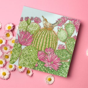 May include: A cross-stitch pattern featuring a cactus with pink flowers and a small bird perched on top. The cactus is surrounded by other cacti and flowers. The background is a light blue color.