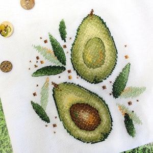 May include: Cross-stitch embroidery design featuring two avocado halves with green leaves and brown seeds on a white background.
