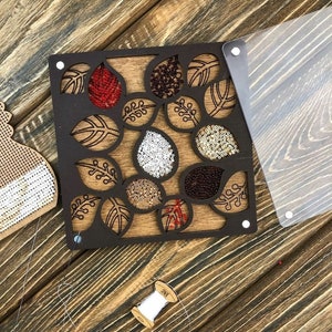 May include: A wooden craft kit with a clear plastic cover. The kit features a design of leaves with cutouts for beads. The kit includes a spool of white thread and a variety of colored beads.