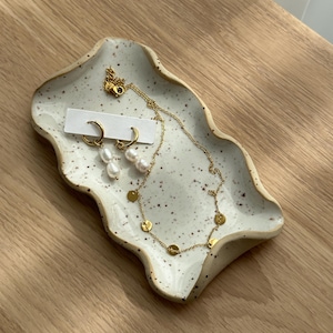 Könnte beinhalten: Eine weiße Keramik-Schale mit gewelltem Rand, auf der eine goldene Kettenkette mit kleinen goldenen Kreisen und ein Paar goldene Creolen mit Perlenakzenten liegen.