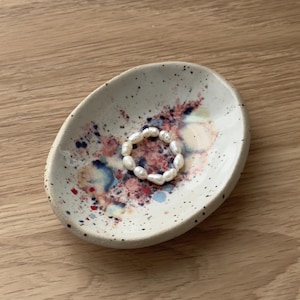 Könnte beinhalten: Ovale Schmuckschale aus Keramik mit einer gesprenkelten, cremefarbenen Oberfläche und farbenfrohen abstrakten Farbspritzern in Rot, Blau und Rosa. Ein Ring aus weißen Perlen liegt in der Schale.