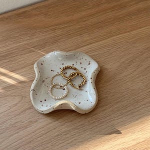 Könnte beinhalten: Eine kleine, cremefarbene Keramik-Schmuckschale mit einem gesprenkelten Muster. Die Schale enthält zwei goldfarbene Creolen und einen kleineren Ring mit weißen Perlen. Die Schale hat eine einzigartige, organische Form.