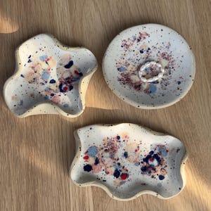 Könnte beinhalten: Drei gesprenkelte Keramik-Schmuckschalen in organischen Formen. Die Schalen sind cremefarben mit farbenfrohen Spritzern in Rot, Blau und Lila. Eine Schale ist rund, die anderen haben unregelmäßige Kanten. Ein Ring liegt in der runden Schale.