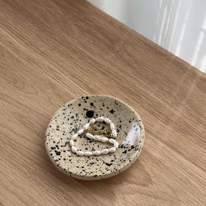 Könnte beinhalten: Eine kleine, gesprenkelte beige Keramik Schale mit schwarzer Sprühfarbe. Eine Perlenkette liegt in der Schale.