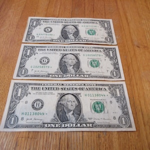 May include: Three United States one-dollar bills are displayed on a wooden surface. Each bill features a portrait of George Washington and the text "ONE DOLLAR". The bills are in shades of green and black.