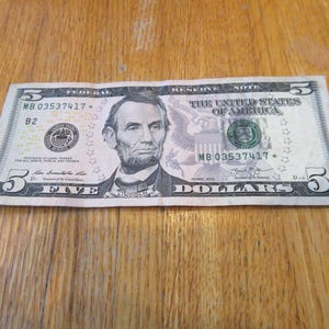 May include: A five-dollar bill featuring a portrait of Abraham Lincoln. The bill displays the text "FEDERAL RESERVE NOTE" and "THE UNITED STATES OF AMERICA". The number "5" is visible in multiple locations.