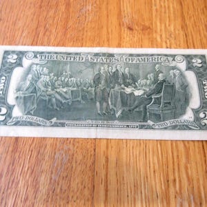May include: A two-dollar bill featuring a detailed engraving of the signing of the Declaration of Independence. The bill is green and white, with the text "The United States of America" at the top. The denomination "TWO" is visible.