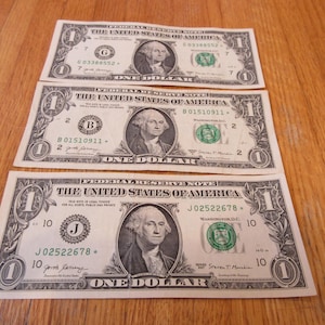 May include: Three one-dollar bills are displayed on a wooden surface. Each bill features a portrait of George Washington and the text "The United States of America." The bills are in good condition, with clear details.