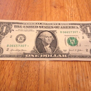 May include: A United States one-dollar bill, featuring a portrait of George Washington. The bill displays the text "The United States of America" and "One Dollar". The bill is green and black.